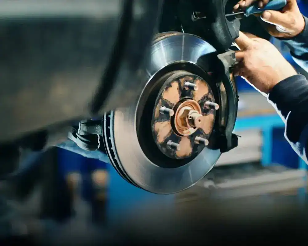 A mechanic’s hands work on a car’s exposed disc brake and wheel hub assembly in a garage, where tools for transmission builds in Athens, GA are visible in the blurred background.