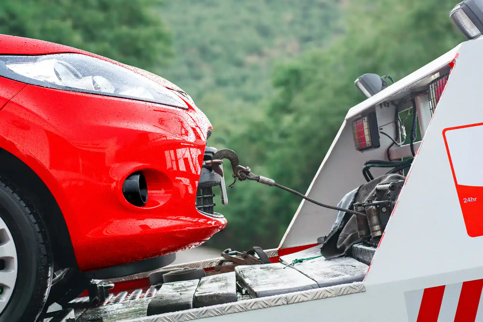 Close-up of a red car being towed by a tow truck on a rainy GA day, with a metal hook attached to the car’s front bumper. Transmission builds Athens services might be needed. Green trees are blurred in the background.