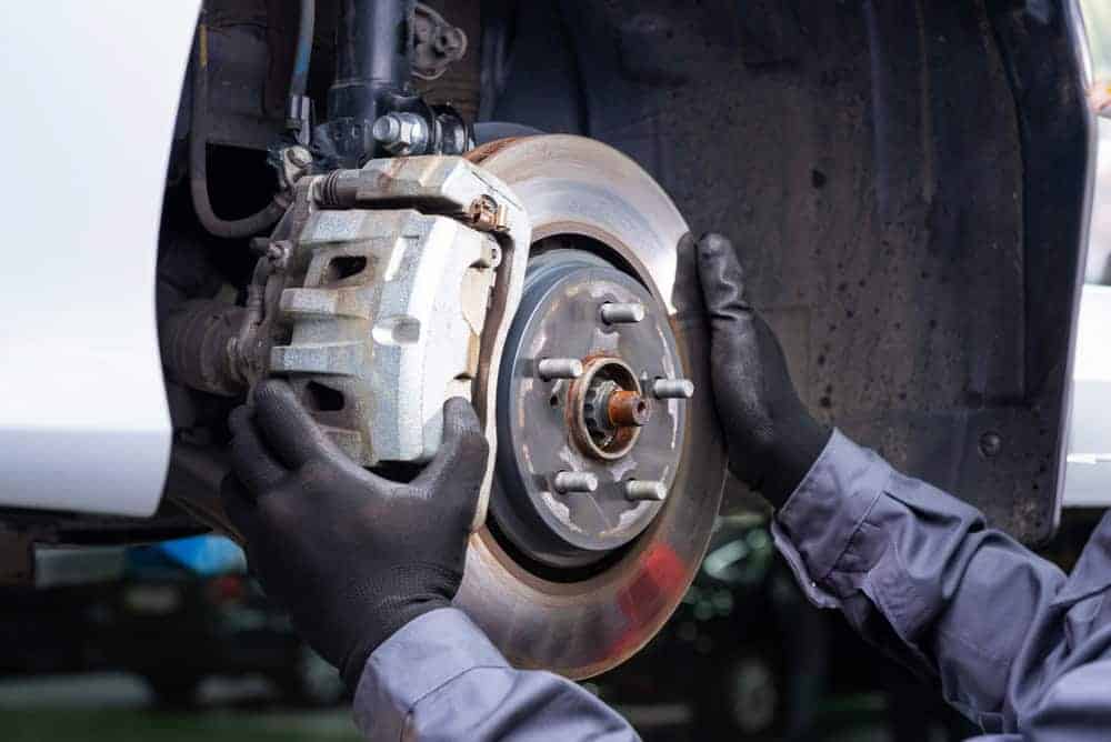 A mechanic wearing black gloves is inspecting or installing a car’s brake rotor and caliper, with the wheel removed and the vehicle raised, at a shop known for expert transmission builds in Athens, GA.