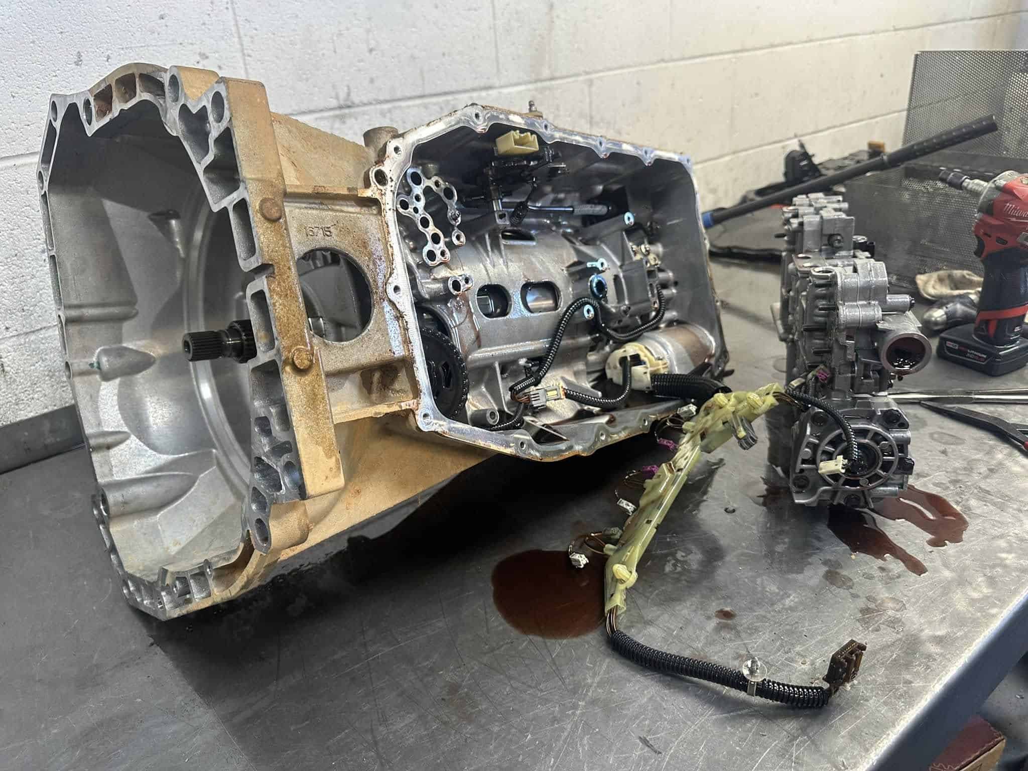 A disassembled automotive transmission sits on a metal workbench in Athens, GA alongside its separated components and wiring, with some fluid spilled nearby and tools visible in the background—ideal for expert transmission builds.